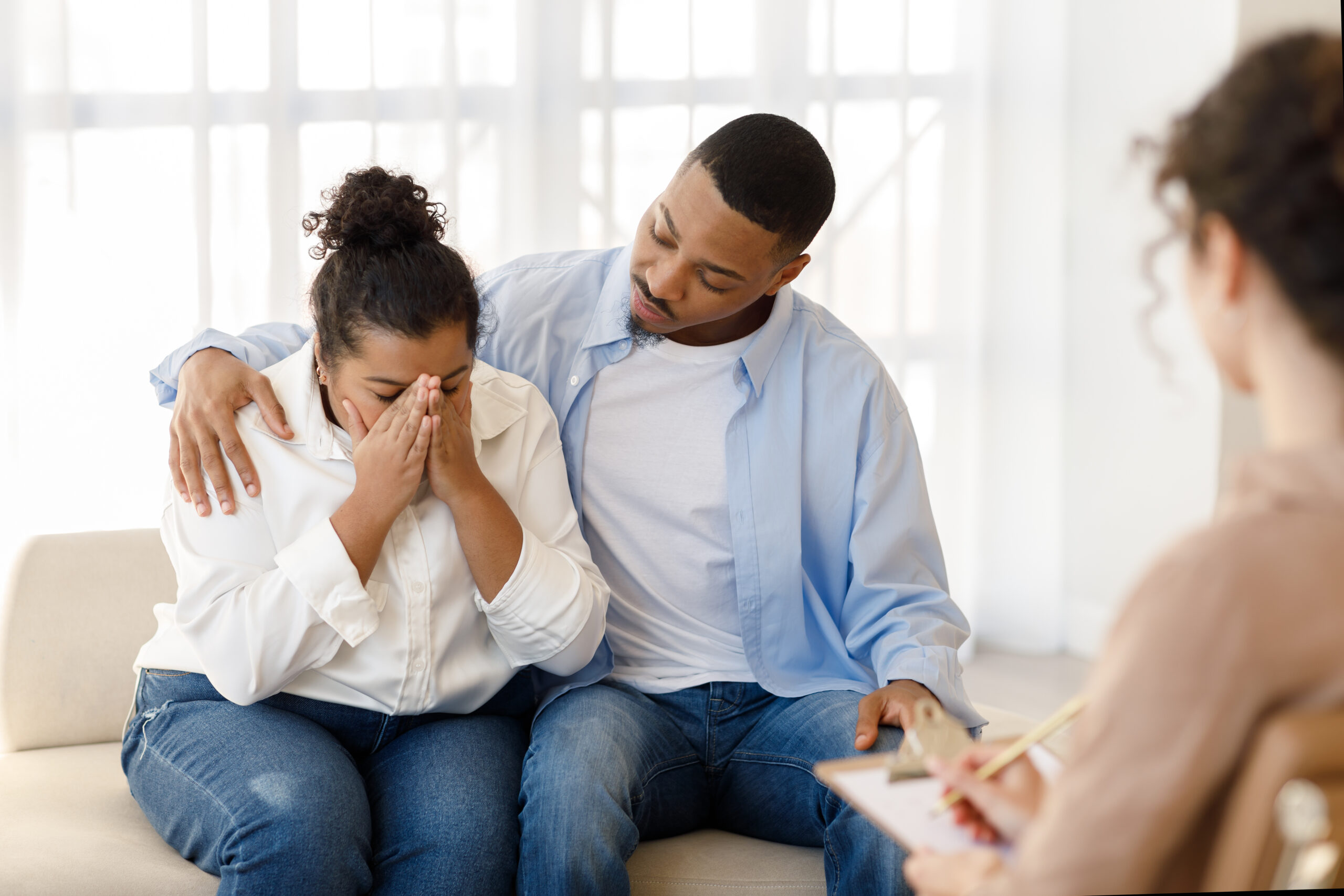 Loving attentive young black guy hugging comforting his crying spouse girlfriend suffering from depression while having family therapy with woman psychologist at counselor office